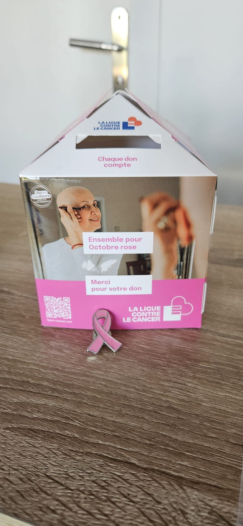 La tirelire de la Ligue contre le cancer posée au salon, avec un pin ruban rose devant
